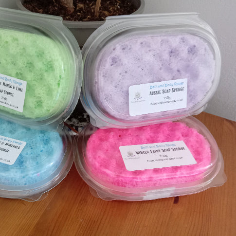 Three clear plastic containers with soap infused sponges inside. The sponges are in a green, blue, and pink color and each container has a label with the product name and description.