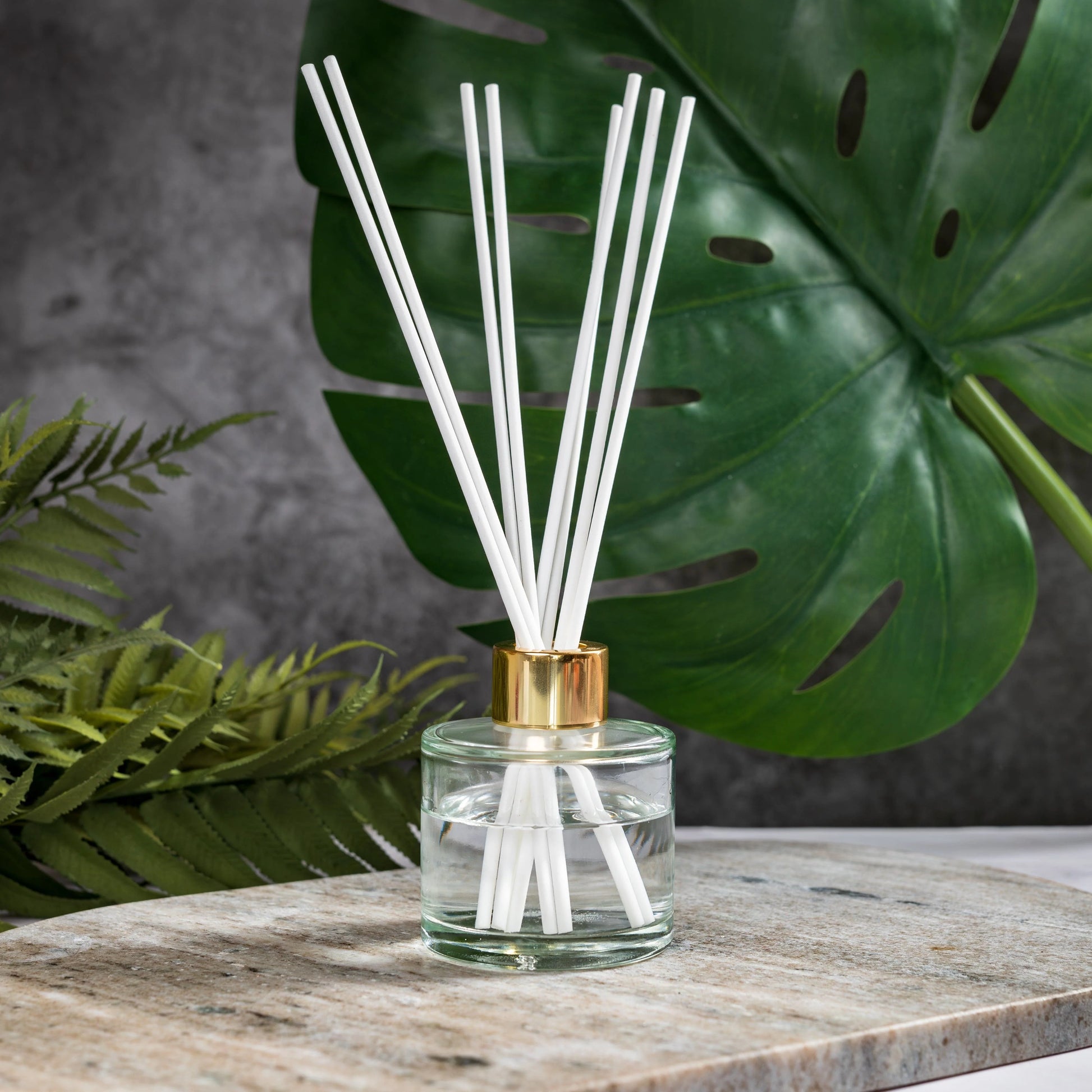 Reed diffuser with white sticks in a clear bottle with gold cap on a wooden surface with green leaves in the background.