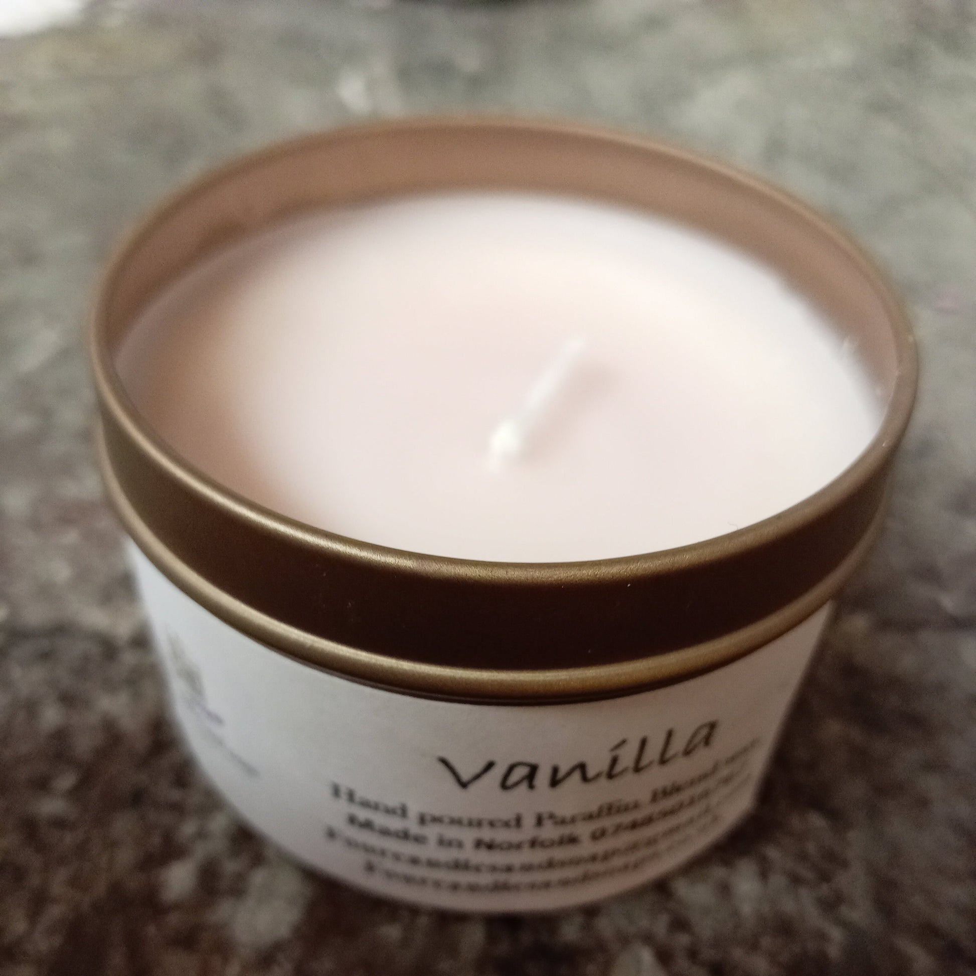 A tin candle with a clear label that reads 'Vanilla hand-poured candle' with a lit wick, placed on a surface with a marble-like texture.