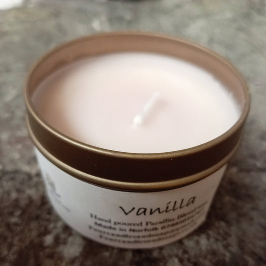 A tin candle with a clear label that reads 'Vanilla hand-poured candle' with a lit wick, placed on a surface with a marble-like texture.