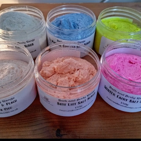 Four jars of Positive Energy Salt Scrub in different colors, arranged on a wooden surface. four candl;es and soaps